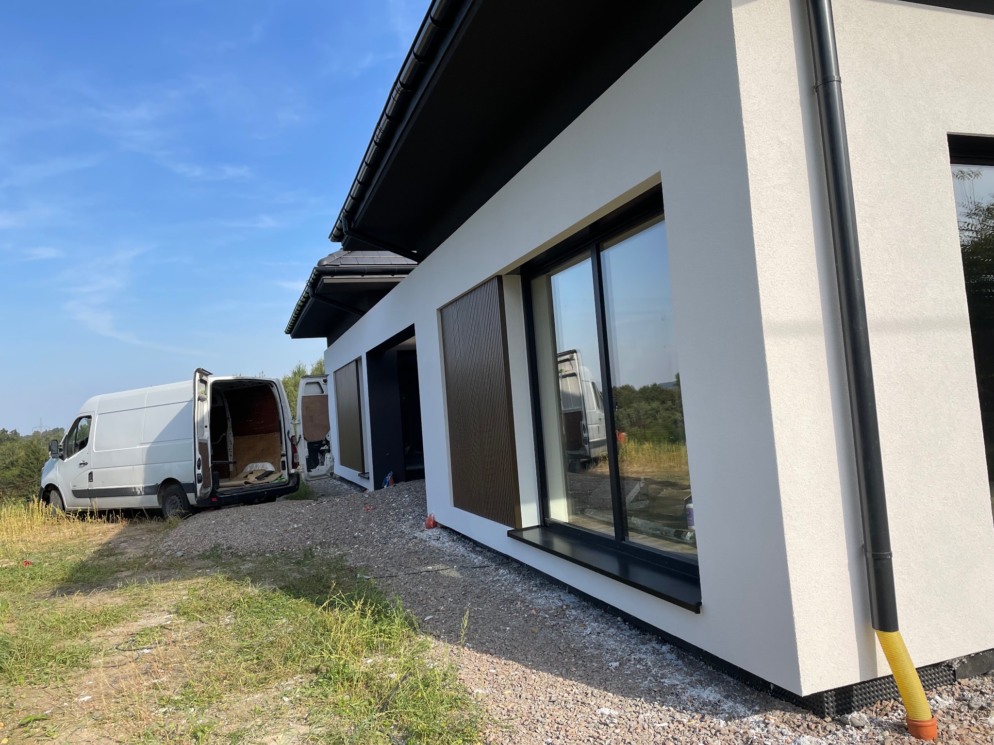 Nowoczesny dom z białą elewacją i czarnymi detalami, widoczny fragment elewacji z oknem, rynną i panelem dekoracyjnym, na pierwszym planie gruz i trawa, w tle biały bus z otwartymi drzwiami.