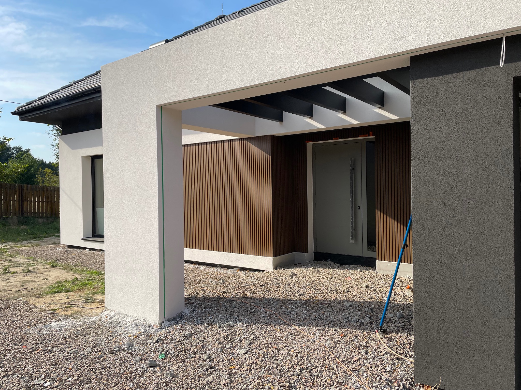 Nowoczesny dom z białą elewacją o fakturze baranka, drewnianą okładziną pionową pod zadaszeniem i antracytowym akcentem, widoczne drobne kamienie wokół budynku.