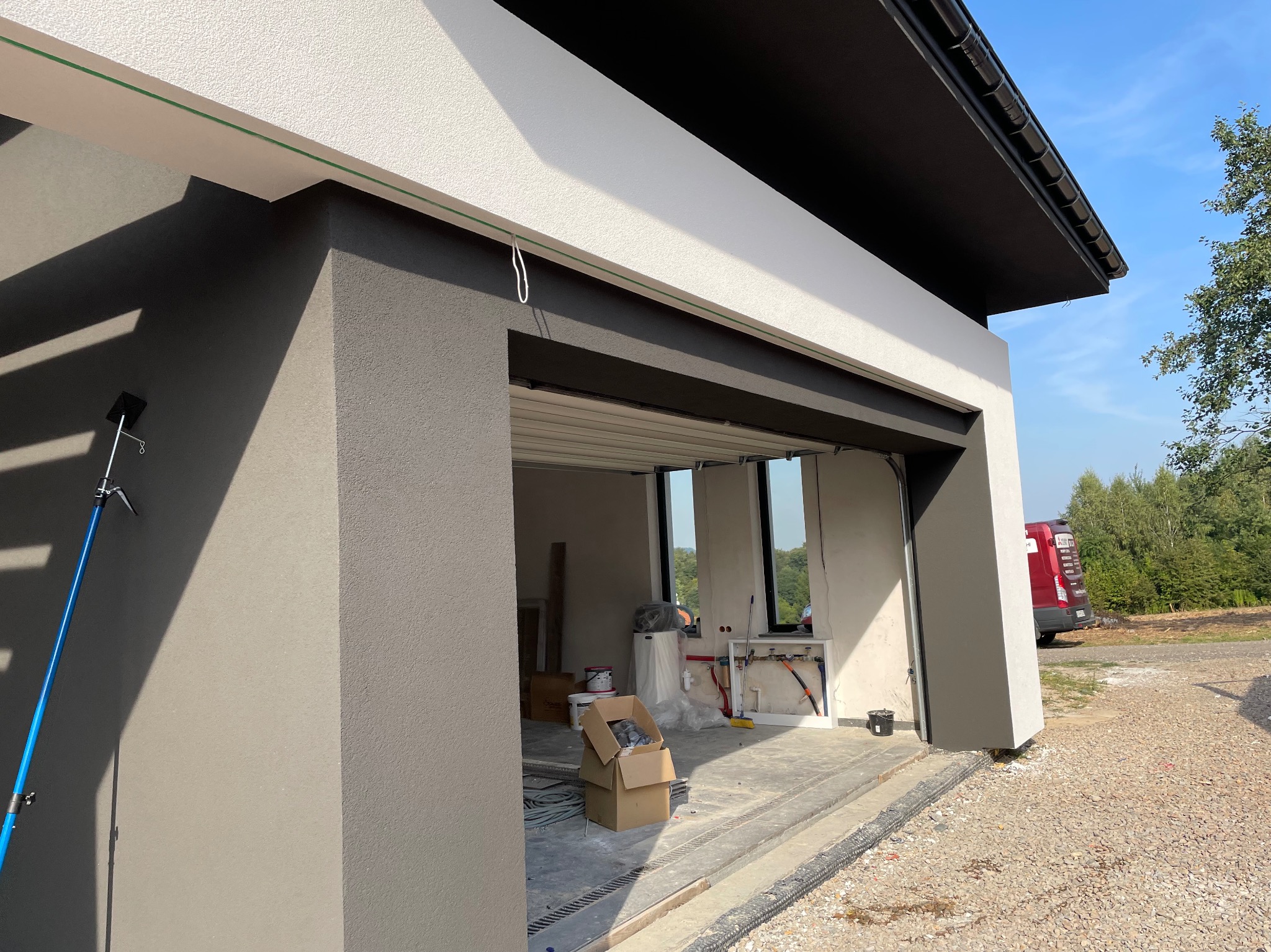 Nowoczesny garaż z elewacją w odcieniach szarości i bieli, widok z zewnątrz na wnętrze w trakcie wykańczania, z widocznymi instalacjami i narzędziami, na zewnątrz zaparkowany czerwony samochód.