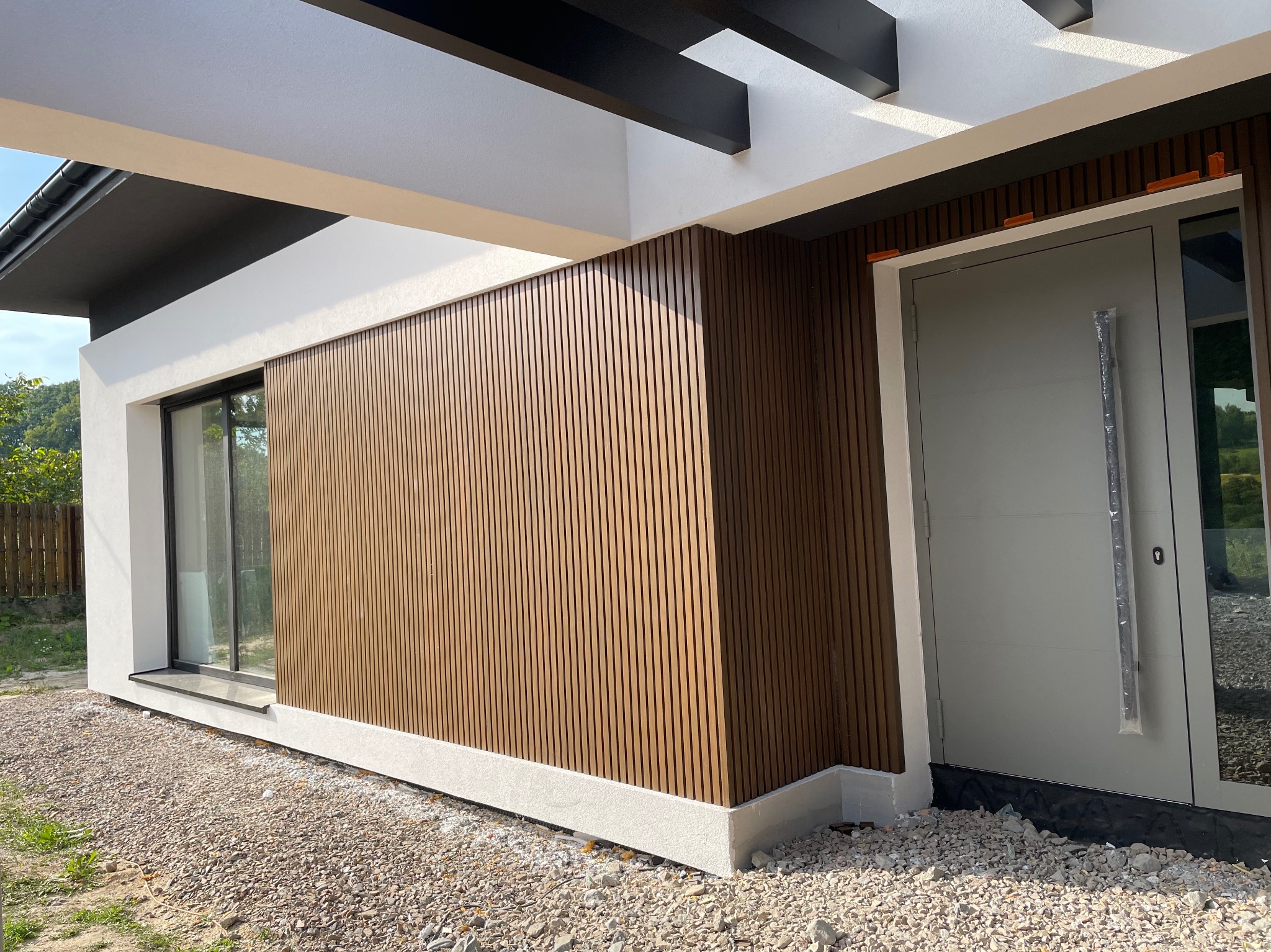 Nowoczesny dom z elewacją z pionowych, drewnopodobnych paneli, białymi ramami okiennymi i drzwiami wejściowymi, otoczony żwirem.