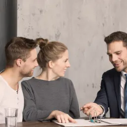 Para rozmawia z doradcą finansowym nad dokumentami na drewnianym stole, obok szklanki wody i roślin w skrzynce