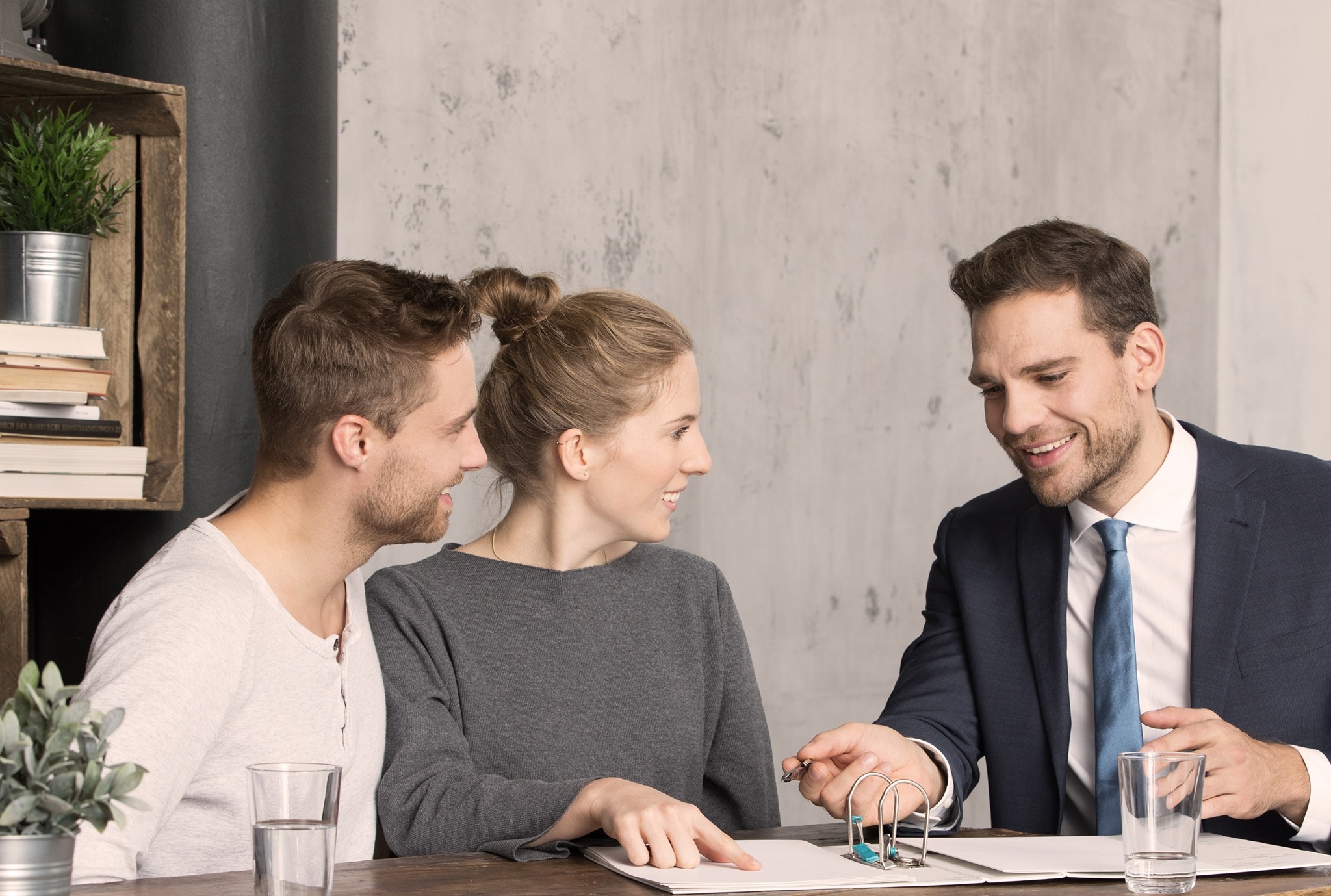 Para rozmawia z doradcą finansowym nad dokumentami na drewnianym stole, obok szklanki wody i roślin w skrzynce