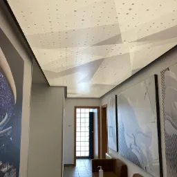 Nowoczesny sufit napinany z geometrycznym wzorem kropek w jasnym korytarzu biurowym z minimalistycznymi meblami i abstrakcyjnymi obrazami na ścianach.