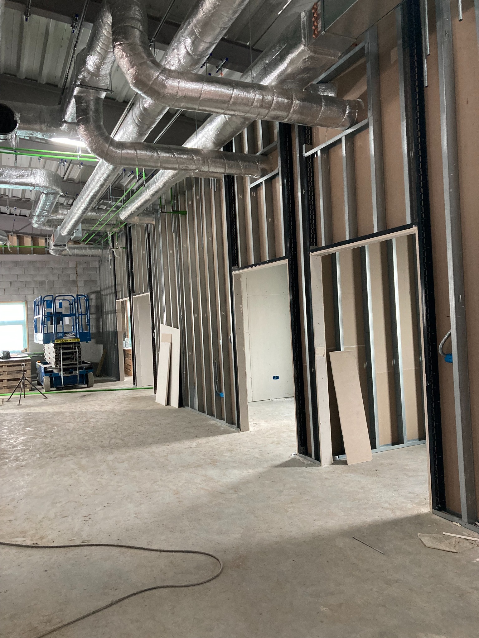Wnętrze w trakcie prac wykończeniowych: widoczne metalowe profile ścian działowych, instalacja wentylacyjna pod sufitem, podnośnik nożycowy w tle przy oknie.
