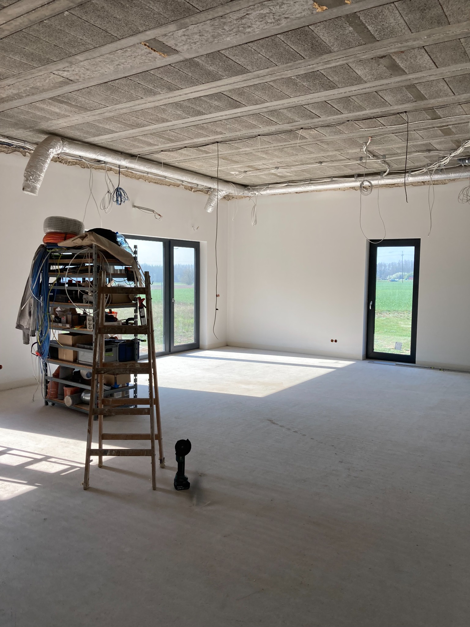 Wnętrze w trakcie remontu: surowy betonowy sufit z widocznymi instalacjami, białe ściany, okna z widokiem na zieleń, drabina z narzędziami i materiałami budowlanymi, szlifierka kątowa przy drabinie.
