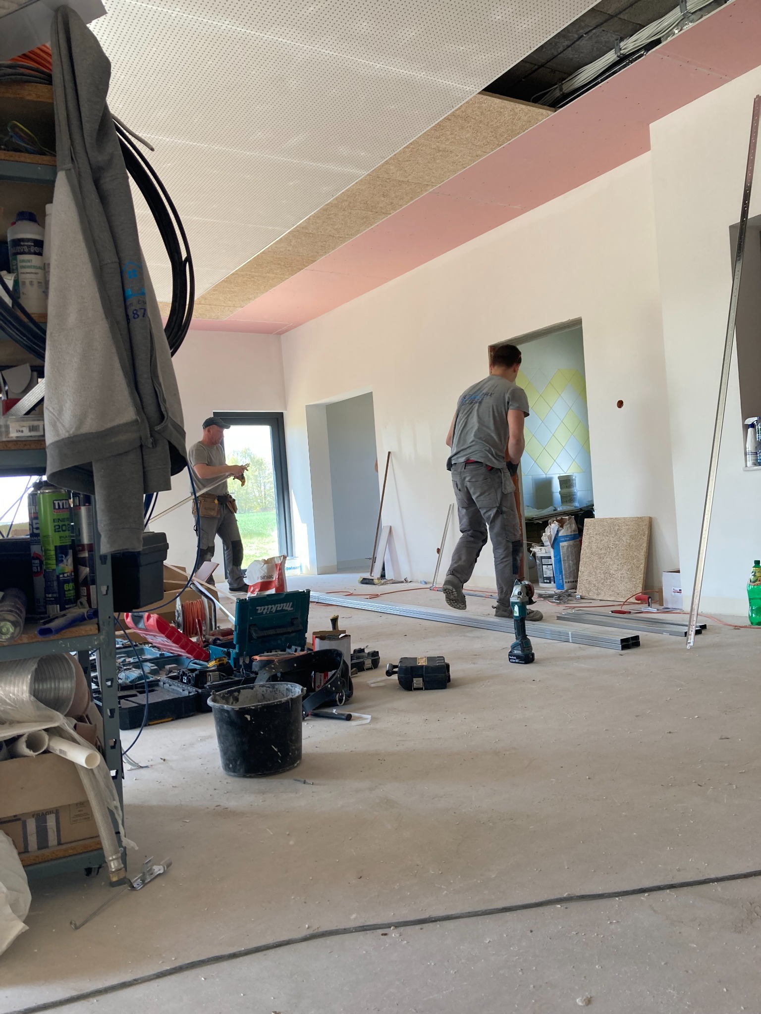 Wnętrze w trakcie prac wykończeniowych: dwóch mężczyzn pracuje nad montażem płyt kartonowo-gipsowych na suficie, widoczne narzędzia, profile aluminiowe, surowy beton na podłodze i częściowo odkryta...