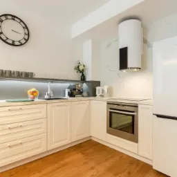 Jasna kuchnia z drewnianą podłogą, białymi szafkami, owocami na blacie i dużym zegarem na ścianie.