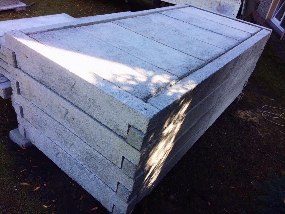 Stos szarych, betonowych prefabrykatów z widocznymi łączeniami typu wpust-wypust, ułożonych na ziemi w słoneczny dzień.