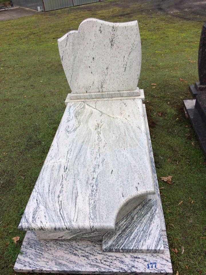 Jasny, gotowy nagrobek z białego granitu z szarymi żyłkami, ustawiony na trawie.