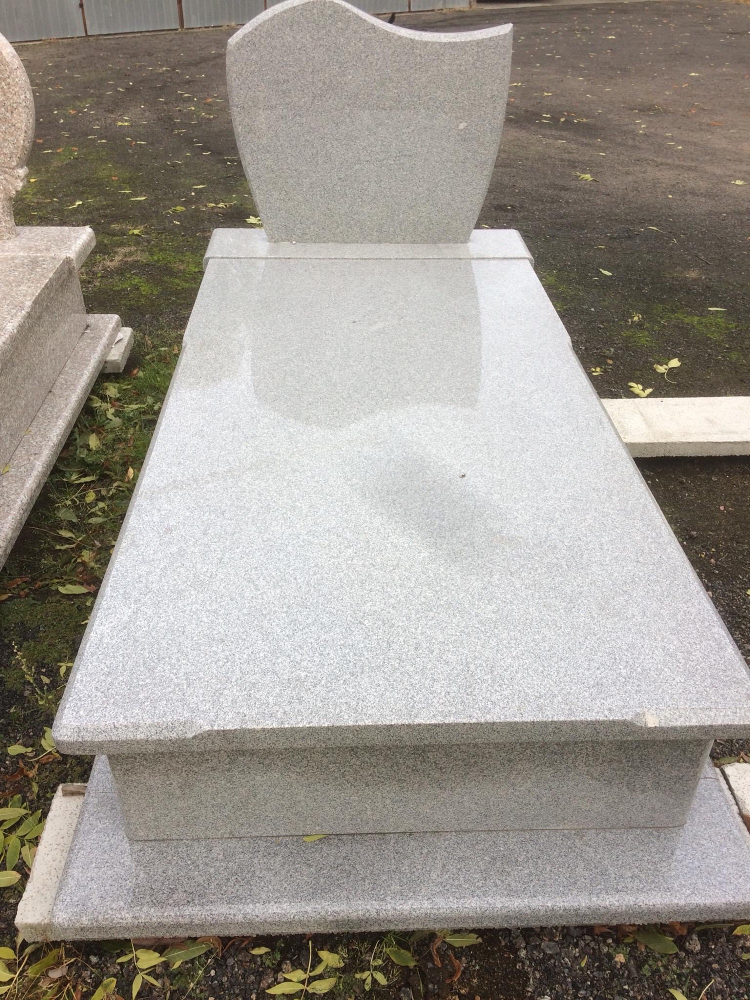 Szary, granitowy pomnik z płytą poziomą i pionową tablicą, ustawiony na zewnątrz na ziemi z liśćmi i mchem.