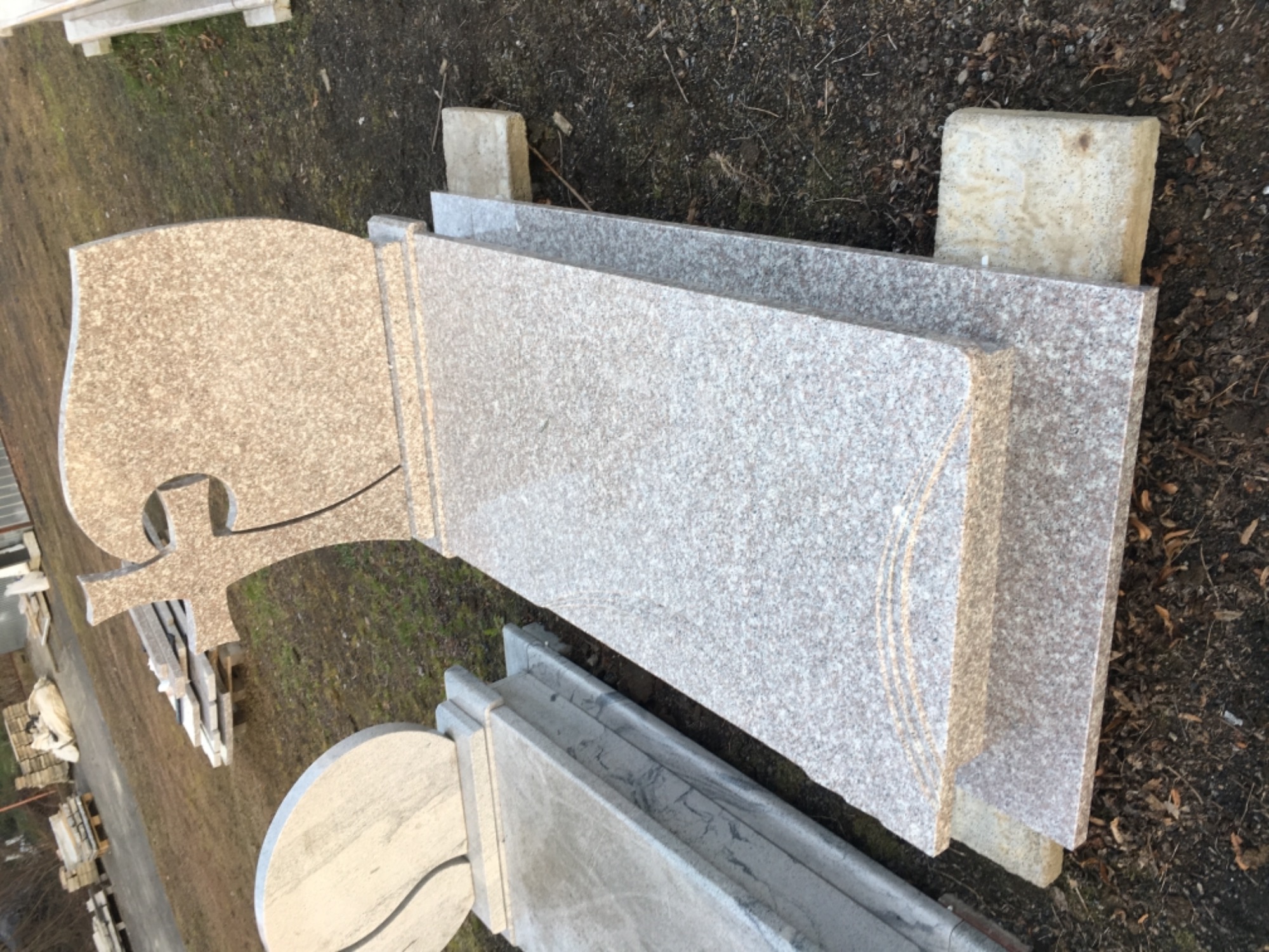 Elementy nagrobne z jasnego granitu, w tym płyta z wyciętym krzyżem i półokrągłym motywem, składowane na betonowych podstawkach na zewnątrz.