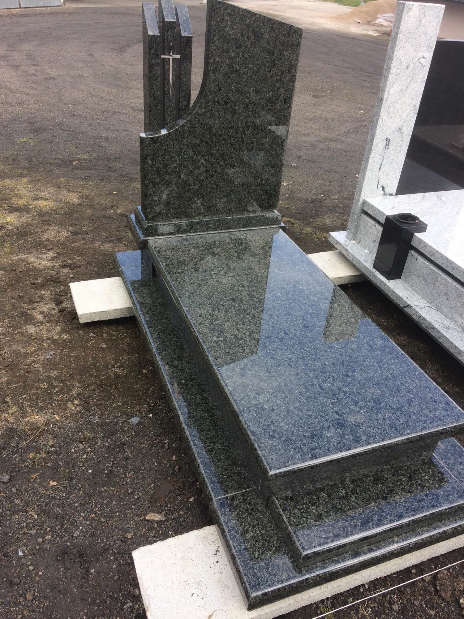 Granitowy pomnik z krzyżem, w odcieniach szarości i zieleni, z polerowaną powierzchnią odbijającą otoczenie, ustawiony na betonowych blokach.
