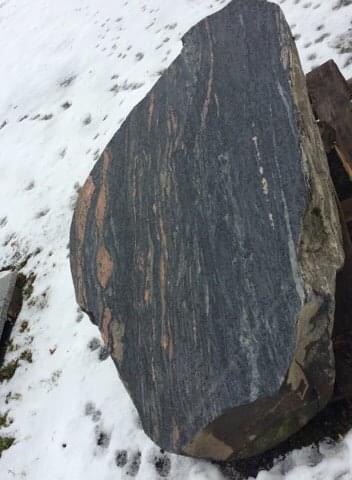 Duży, surowy blok ciemnego kamienia z wyraźnymi, podłużnymi, czerwono-brązowymi żyłkami, leżący na śniegu.
