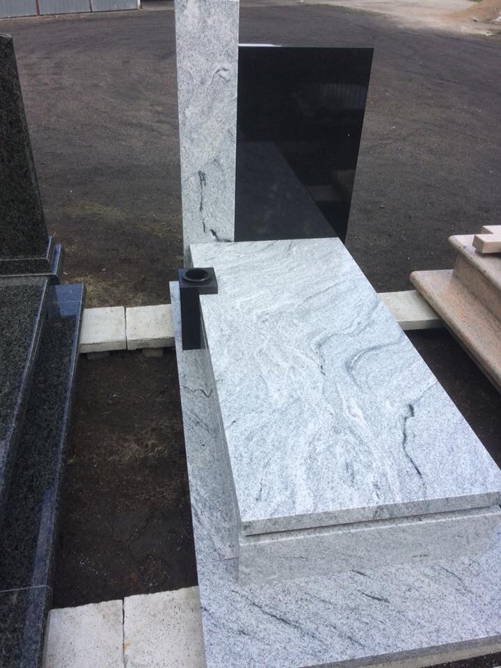 Jasnoszary pomnik z czarnym elementem, widoczny z góry, ustawiony obok innych nagrobków na placu kamieniarskim.