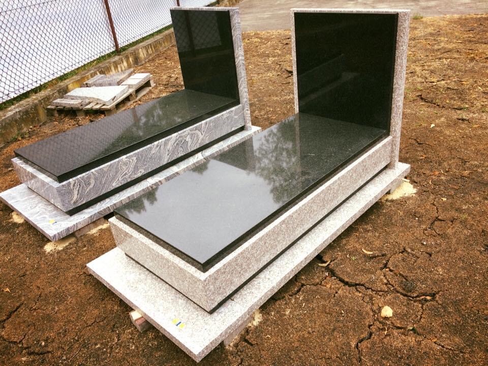 Dwa gotowe, czarne i szare pomniki z kamienia stojące na brązowej ziemi, widoczne palety w tle.