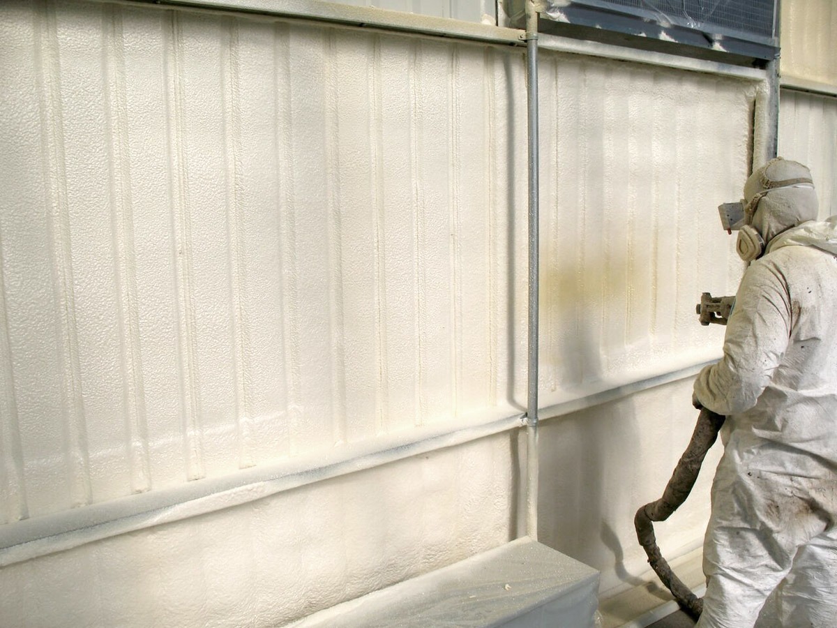 Pracownik w kombinezonie ochronnym natryskuje białą pianę izolacyjną na wewnętrzną ścianę budynku o konstrukcji stalowej.