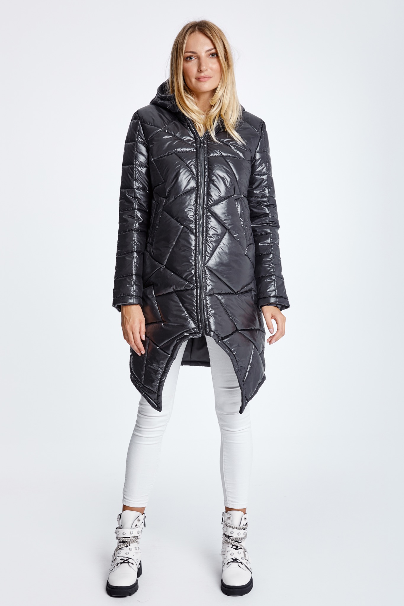 Modelka prezentuje błyszczącą, pikowaną kurtkę z asymetrycznym dołem, w połączeniu z białymi spodniami i butami w stylu rockowym na białym tle.