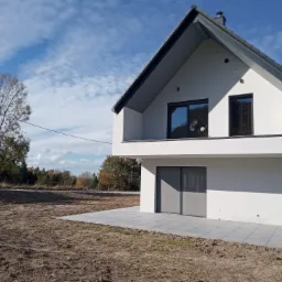 Nowoczesny, dwukondygnacyjny dom z białym tynkiem i szarą stolarką okienną, otoczony przygotowanym terenem pod trawnik i kostką na tarasie, widok na błękitne niebo z chmurami.