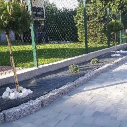 Granitowa obrzeża chodnika z młodymi drzewkami i agrowłókniną, widok na posesję z ogrodzeniem z siatki.