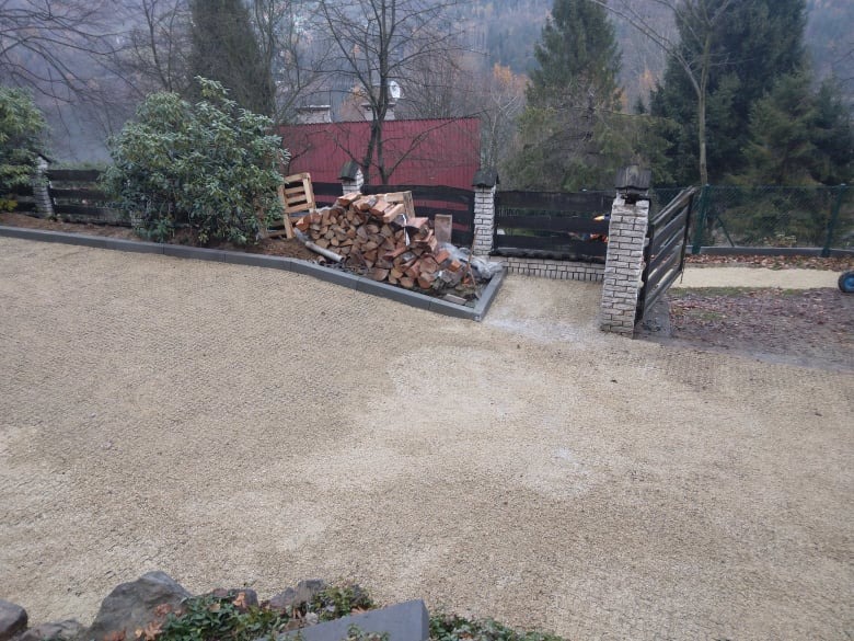 Wykończona żwirowa droga dojazdowa z obrzeżami z kostki, stos drewna przy ogrodzeniu z ceglanymi słupkami, widok na góry w tle.