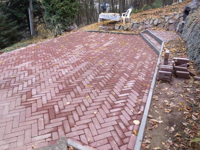 Nowo ułożona czerwona kostka w jodełkę na podjeździe w Bystrej, z widocznymi stosami kostki do wykończenia i schodami z kostki prowadzącymi na wyższy poziom ogrodu.