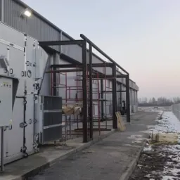 Konstrukcja stalowa w trakcie montażu przy budynku przemysłowym, widoczne rusztowania i elementy przygotowane do spawania, zimowa sceneria z resztkami śniegu.