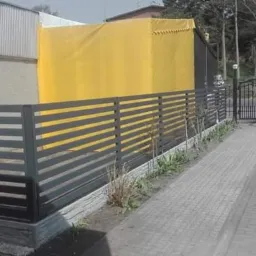 Nowoczesne ogrodzenie posesji z poziomych, szarych profili, z widocznym żółtym zadaszeniem w tle i fragmentem białego samochodu po prawej stronie.