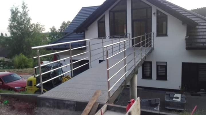 Zewnętrzna rampa dla niepełnosprawnych z metalową konstrukcją i poziomymi barierkami w Kołobrzegu, prowadząca do wejścia do nowoczesnego domu. W tle widoczny częściowo samochód i sprzęt budowlany.