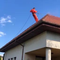 Pracownik w pomarańczowym kombinezonie, zabezpieczony linami, czyści dach z brązowej dachówki za pomocą myjki ciśnieniowej, na tle błękitnego nieba z lekkimi chmurami i fragmentu elewacji budynku.