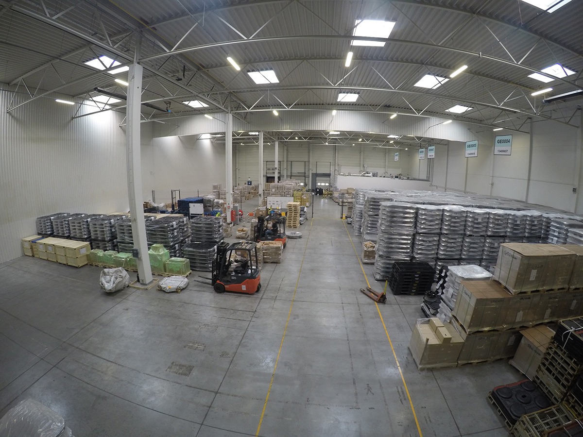 Przestronny magazyn w Gliwicach z wózkiem widłowym, paletami i towarem, oświetlony światłem dziennym wpadającym przez świetliki dachowe. Widoczny podział na strefy za pomocą żółtych linii na podłodze.