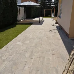 Beżowa kostka brukowa ułożona na posesji, widoczny trawnik, altana z zadaszeniem, huśtawka dla dzieci i wąż ogrodowy