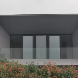 Nowoczesny budynek z szarą elewacją, trzema dużymi oknami balkonowymi za szklaną balustradą, otoczony kwitnącymi czerwonymi makami.