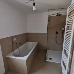 Nowoczesna łazienka z beżowymi płytkami, białą wanną, kabiną prysznicową ze szklaną ścianką i grzejnikiem drabinkowym; surowa żarówka zwisa z sufitu.