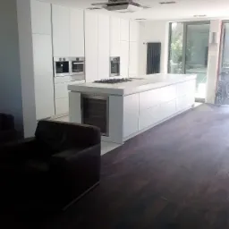 Nowoczesna kuchnia z białymi, minimalistycznymi szafkami, wyspą z płytą grzewczą i wbudowaną chłodziarką do wina, oświetleniem sufitowym i ciemną podłogą, widziana z perspektywy salonu z brązowym...