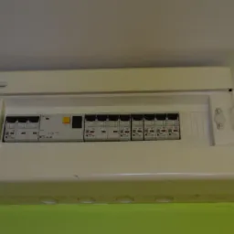 Otwarta skrzynka elektryczna z wyłącznikami nad zieloną ścianą, widoczne logo Electro-Plast.