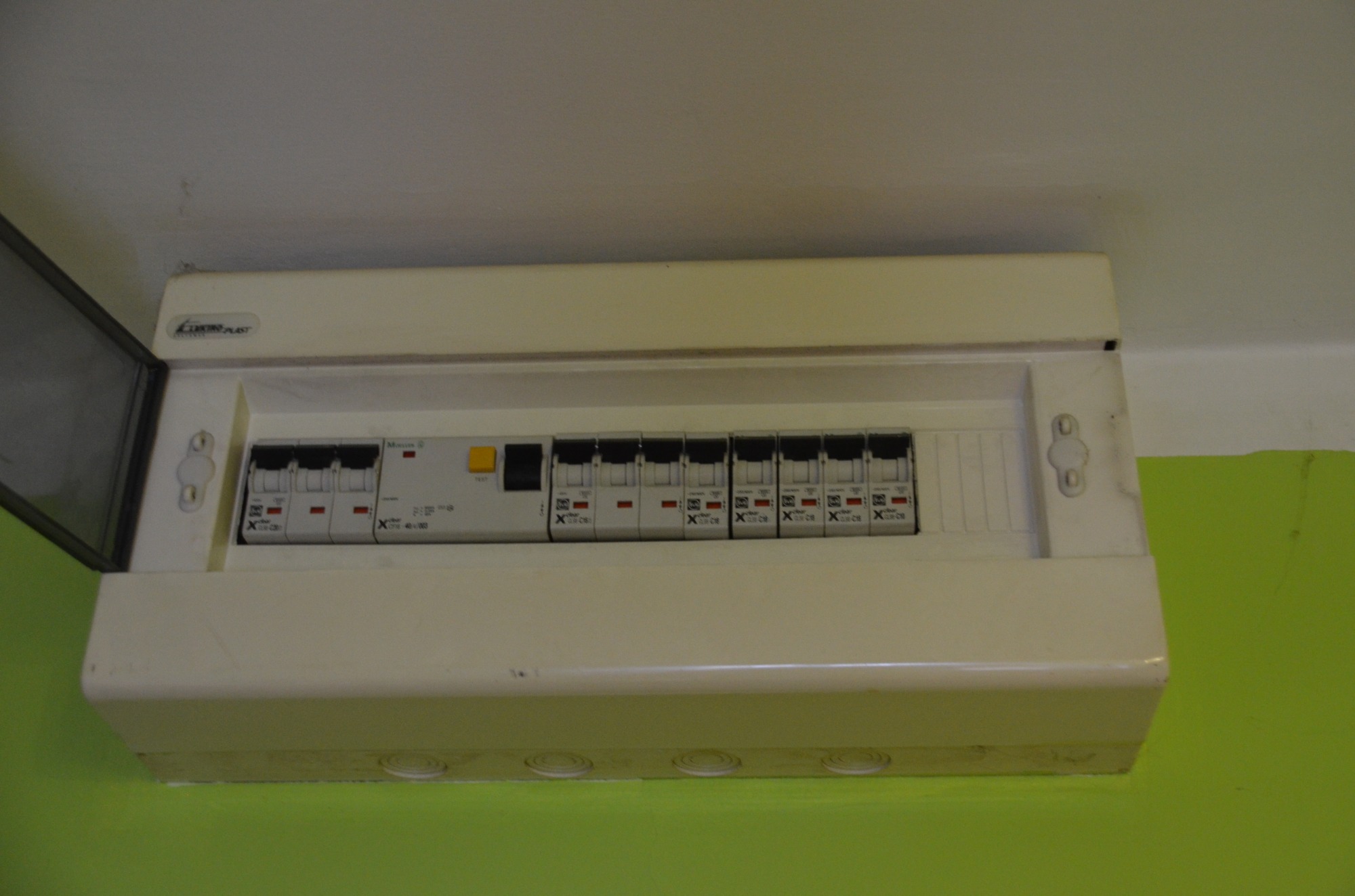 Otwarta skrzynka elektryczna z wyłącznikami nad zieloną ścianą, widoczne logo Electro-Plast.