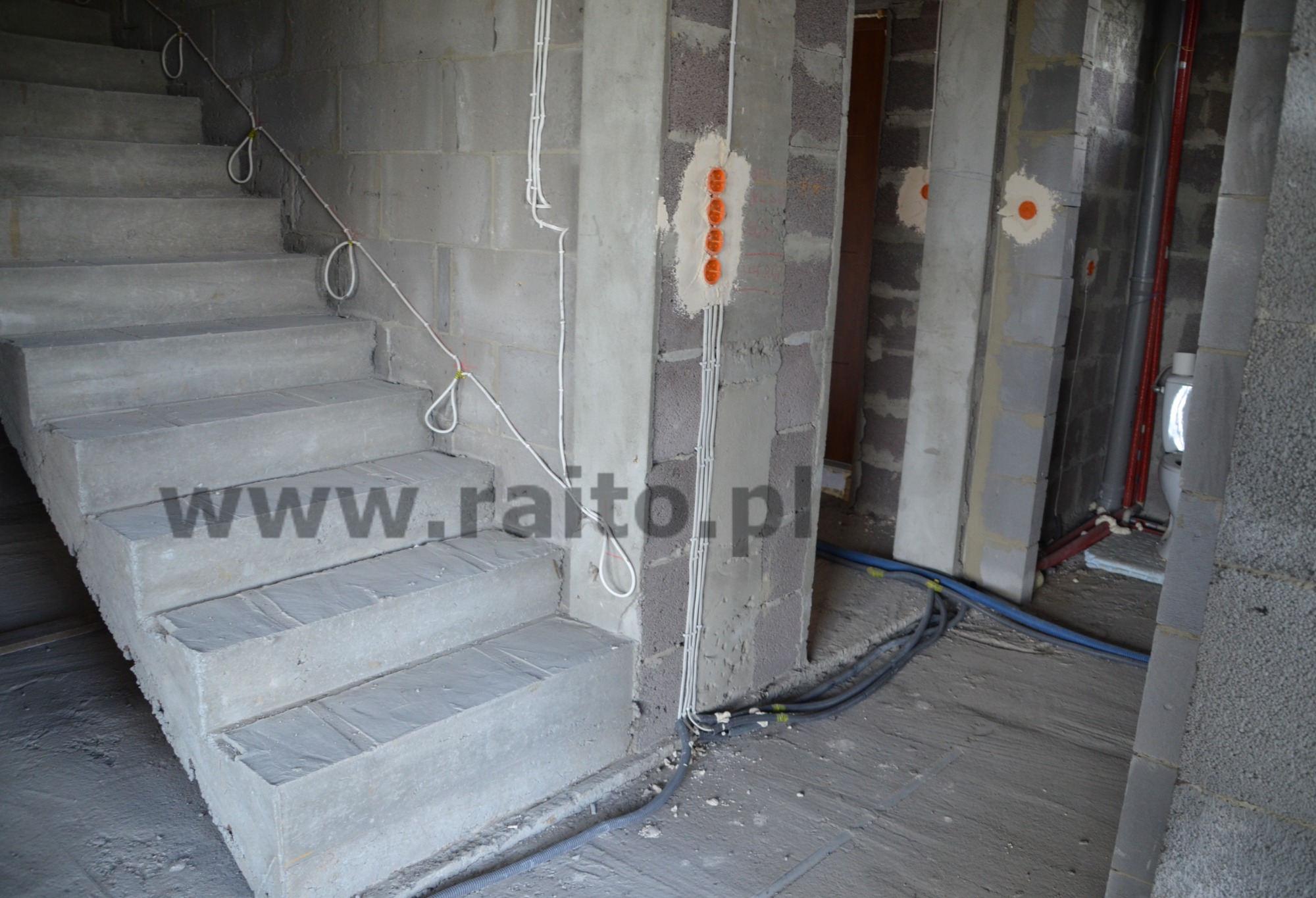 Surowe betonowe schody w trakcie budowy, widoczne kable elektryczne ułożone wzdłuż ściany, puszki elektryczne w ścianie, szare ściany z betonu komórkowego.