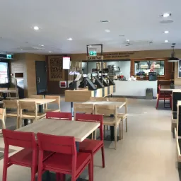 Jasne wnętrze lokalu gastronomicznego z drewnianymi stołami i krzesłami, czerwone akcenty na krzesłach, widoczny bar z kasami i ekspozycją napojów, szyld z ofertą menu na ścianie.