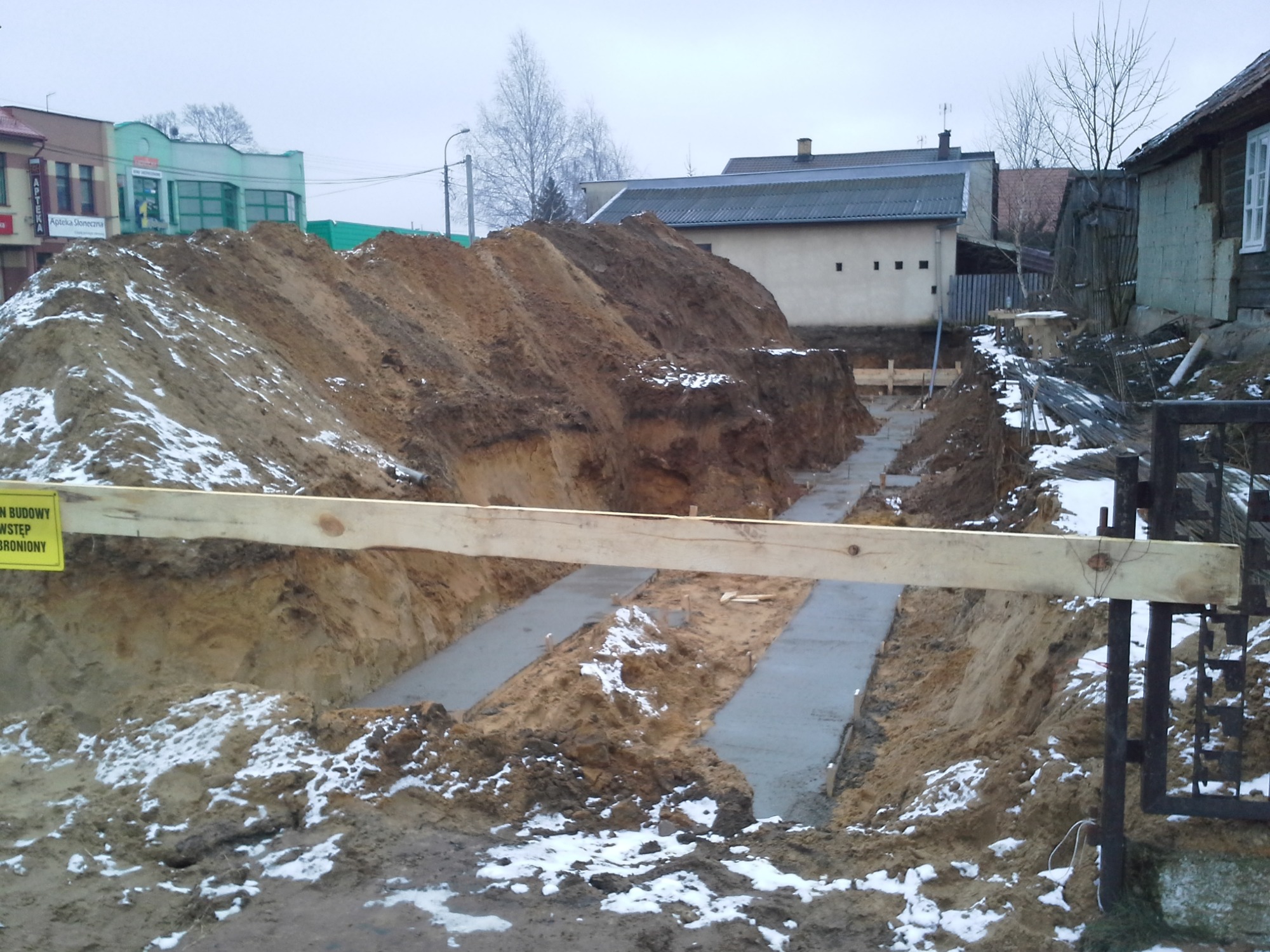 Wykopane fundamenty pod budowę w Białymstoku, widoczne betonowe ławy fundamentowe, ziemia i śnieg, z przodu drewniana belka i fragment ogrodzenia.