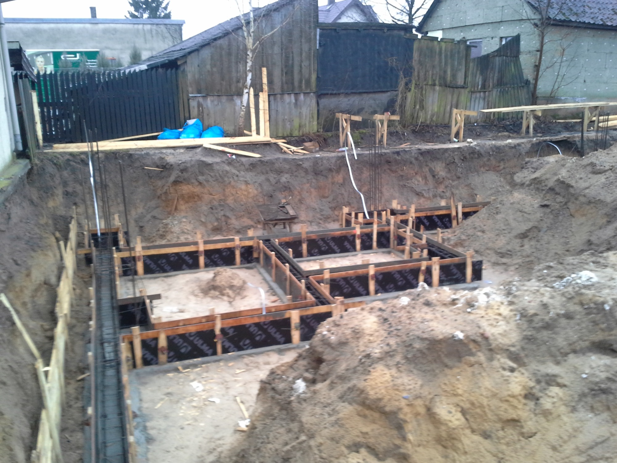 Wykop pod fundamenty z drewnianym szalunkiem i zbrojeniem, widoczne izolacje pionowe, w tle zabudowa jednorodzinna i ogrodzenie z desek
