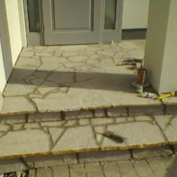 Schody wejściowe wyłożone nieregularnymi płytami kamiennymi, widoczne narzędzia i pozostałości po pracy, fragment kolumny i drzwi wejściowych.