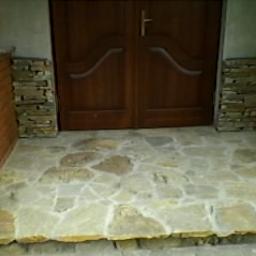 Jerzy Ciesielka Wykonywanie Kominków i Ogrodzeń z Kamienia - Kamienne wykończenie wejścia do domu z nieregularnych płyt piaskowca, otoczone ceglanymi i kamiennymi elementami dekoracyjnymi, drewniane drzwi wejściowe w tle.