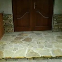 Kamienne wykończenie wejścia do domu z nieregularnych płyt piaskowca, otoczone ceglanymi i kamiennymi elementami dekoracyjnymi, drewniane drzwi wejściowe w tle.