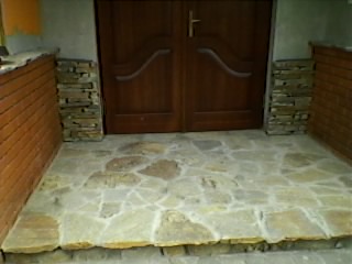 Kamienne wykończenie wejścia do domu z nieregularnych płyt piaskowca, otoczone ceglanymi i kamiennymi elementami dekoracyjnymi, drewniane drzwi wejściowe w tle.