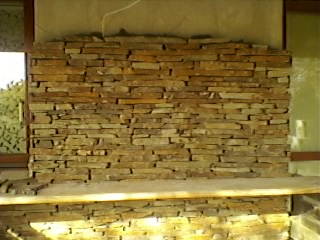 Ściana wykończona dekoracyjnym kamieniem w odcieniach brązu i beżu, ułożonym w poziome pasy, widoczne niedoskonałości i resztki zaprawy, surowy stan wykończenia.