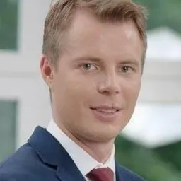 Portret mężczyzny w garniturze z czerwoną koszulą i krawatem, uśmiechający się, z krótkimi blond włosami, na rozmytym zielonym tle.