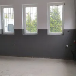 Wnętrze pomieszczenia w trakcie prac wykończeniowych, ściana dwukolorowa, szara w dolnej części, biała w górnej, trzy okna z kratami, podłoga wyłożona płytkami, widoczna brama garażowa i materiały...