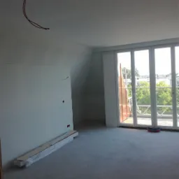 Pokój w trakcie remontu z przygotowanymi ścianami, widoczne przewody elektryczne na suficie, okno balkonowe z widokiem na zieleń, oraz paczka płyt przy ścianie.