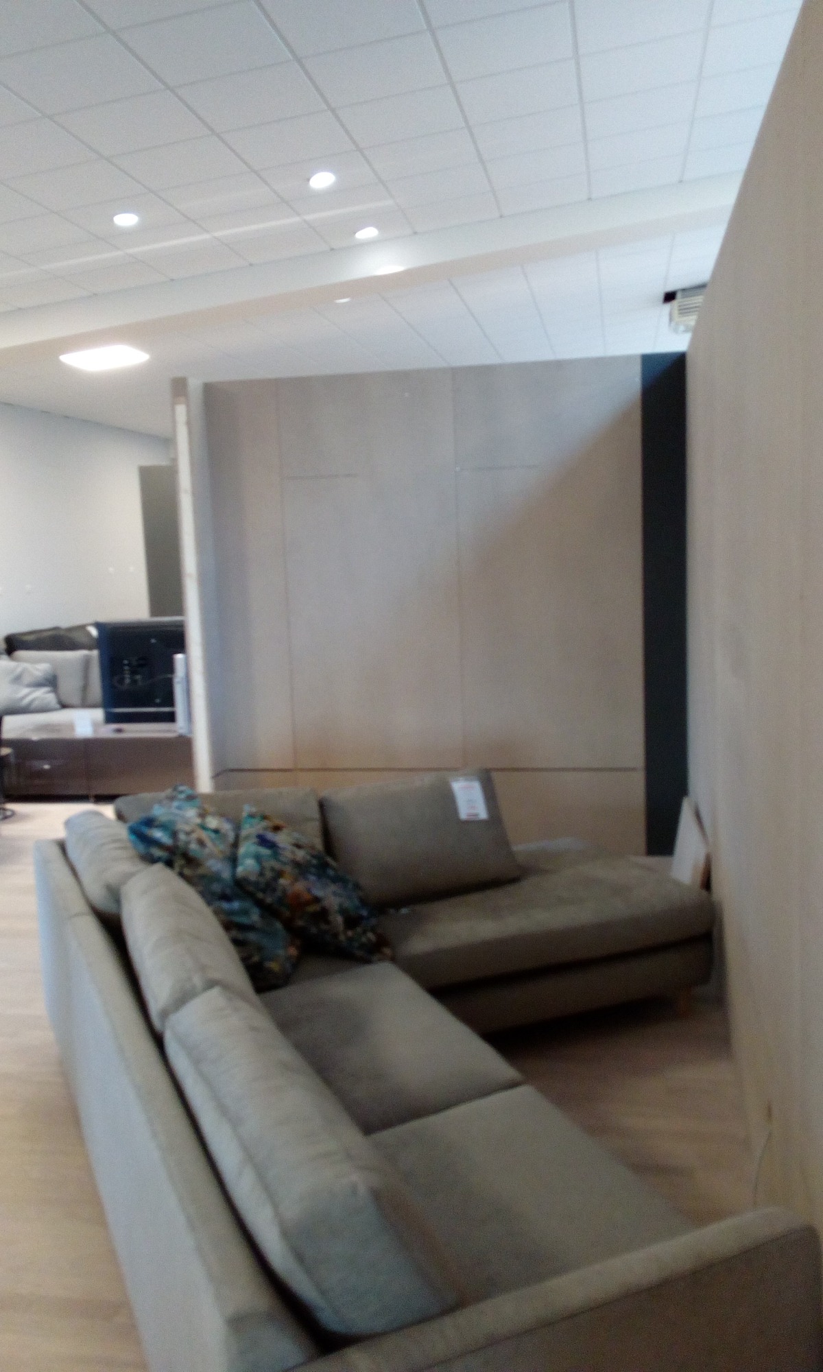 Szary narożnik z poduszkami w odcieniach niebieskiego i brązowego, w tle ściana z płyt karton gips, widoczna z boku, w jasnym pomieszczeniu.