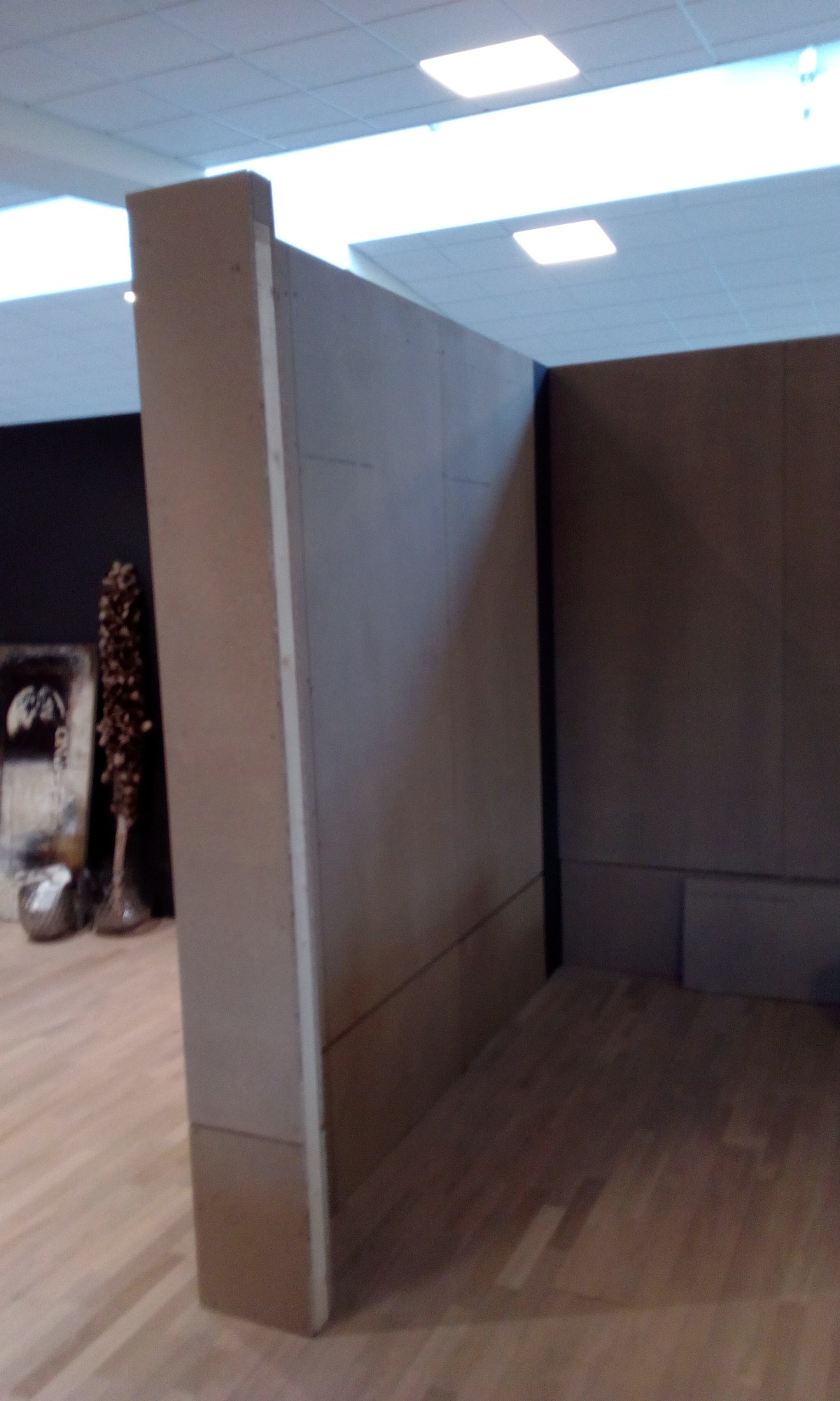 Fragment ściany z płyt kartonowo-gipsowych przed wykończeniem, widoczny surowy montaż i łączenia, na podłodze widoczne drewniane panele, w tle dekoracje.